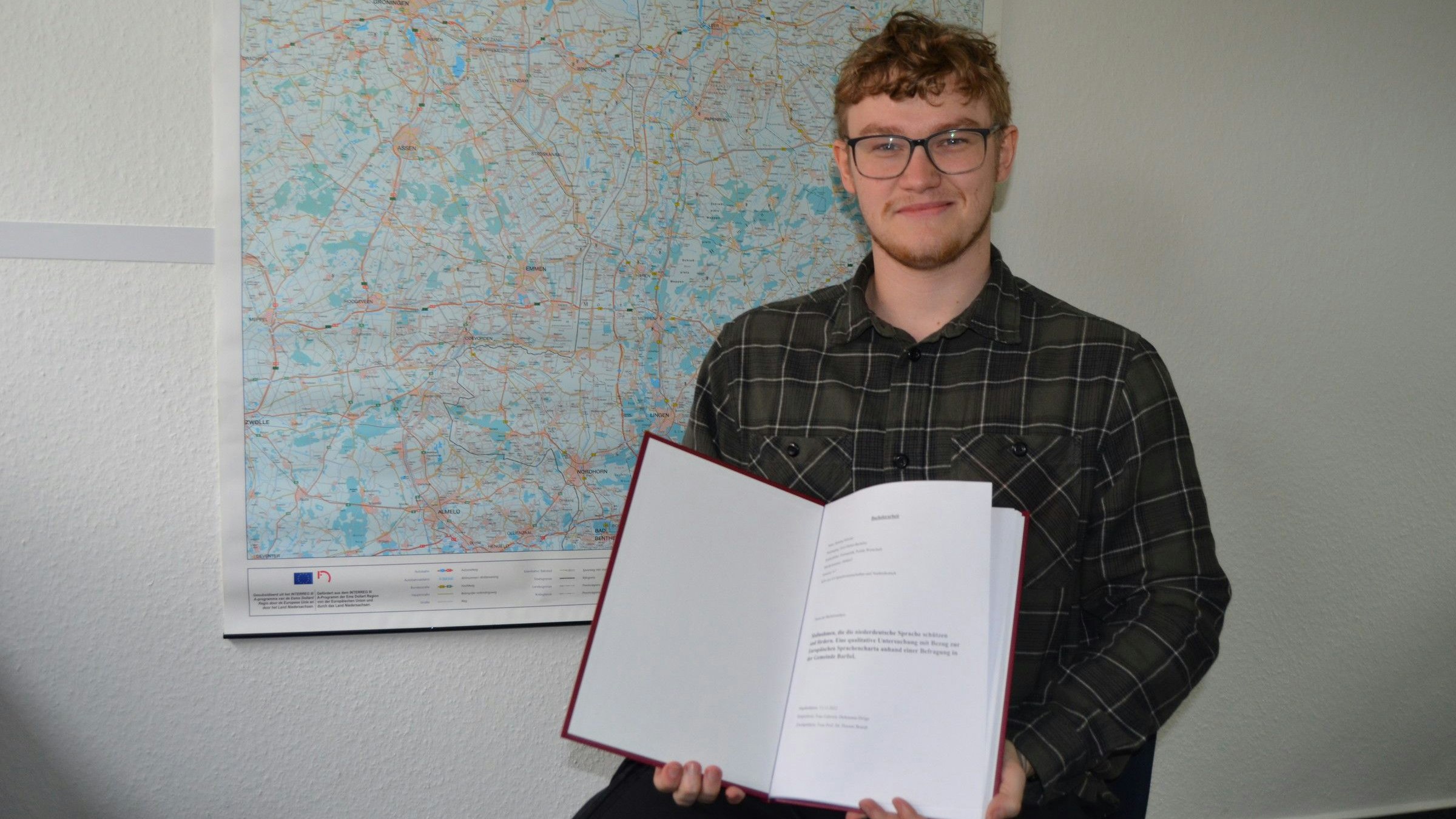 Der 23-jährige Barßeler Henning Schoone mit seiner Bachelorarbeit über das Plattdeutsche vor einer Landkarte Ostfrieslands, wo Platt immer noch sehr verbreitet ist. Foto: Fertig